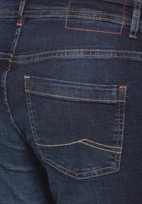 Jean bleu foncé avec une poche arrière dotée de coutures beiges et d'un accent rouge contrastant. La texture en denim est lisse et légèrement délavée.