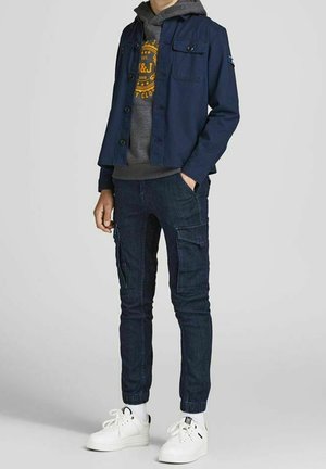 Giacca blu navy con bottoni sopra una felpa grigia con cappuccio, abbinata a jeans cargo blu scuro e sneaker bianche. Presenta tasche sia nei pantaloni che nella giacca.