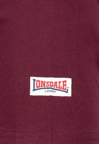 Bordeaux T-shirt met een gladde textuur, voorzien van een wit label met rode en blauwe tekst dat "LONSDALE LONDON" op de onderkant gestikt is.