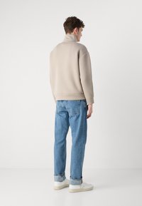 Felpa beige con collo alto, vestibilità rilassata, abbinata a jeans azzurri chiari con orli arrotolati e sneakers bianche. Design minimalista nel complesso.