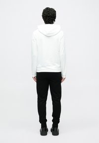 Osoba s kučeravými vlasmi oblečená v bielej mikine s kapucňou, čiernych teplákoch a čiernych teniskách stojaca chrbtom k prostému bielemu pozadiu.