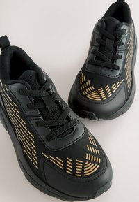 Schwarze Sportschuhe mit strukturiertem Obermaterial, das goldfarbene Akzente und eine Mesh-Zunge aufweist. Ausgestattet mit einer gepolsterten Sohle und Schnürverschluss.
