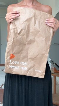 Rudas susiraukšlėjęs popierinis maišelis su baltais užrašais "Mylėk mane. Dėvėk mane." ir Zalando logotipu. Maišelį laiko asmuo, vilkintis juodą sijoną.