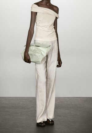 Henkilö, joka pukeutunut olkapäät paljastavaan kermanväriseen toppiin ja löysiin valkoisiin housuihin, pitää kädessään suurta vaaleanvihreää käsilaukkua ja seisoo harmaalla lattialla.