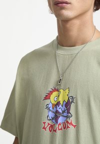 Oliven grøn t-shirt med et tegneserieagtigt grafisk motiv af to fugle på en globe, med røde og gule accenter. Inkluderer en sølvfarvet kæderegulering.