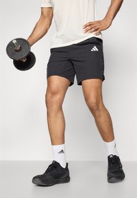 Mustad mustad lühikesed püksid, millel on iseloomulik valge Adidas'i logo. Isik hoiab musta hantlit, kandes musti tossusid ja valgeid sokke.