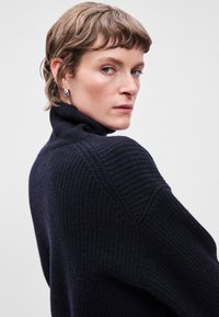 Μπλέ ριπ πλεχτό πουλόβερ με ψηλό λαιμό και χαλαρή γραμμή, που διαθέτει χαμηλωμένους ώμους και υφή. Φοριούνται ασημένια σκουλαρίκια με τρουκ.