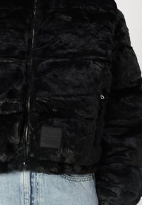 Jachetă scurtă din blană sintetică neagră cu design quiltat, închidere cu fermoar și etichetă cu patch pe față. Textură moale cu accente strălucitoare.