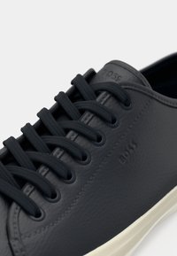 Gros plan d'une basket en cuir noir avec des lacets noirs et le logo BOSS embossé sur le côté, avec une semelle en caoutchouc blanche.