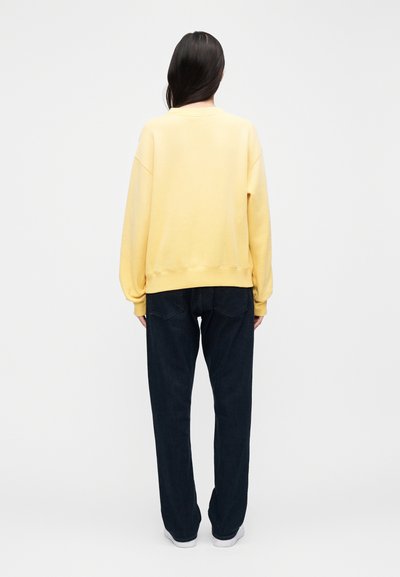 Yellow cotton sweatshirt with dropped shoulders, ribbed cuffs, and hem, paired with dark blue loose-fit jeans and white sneakers.