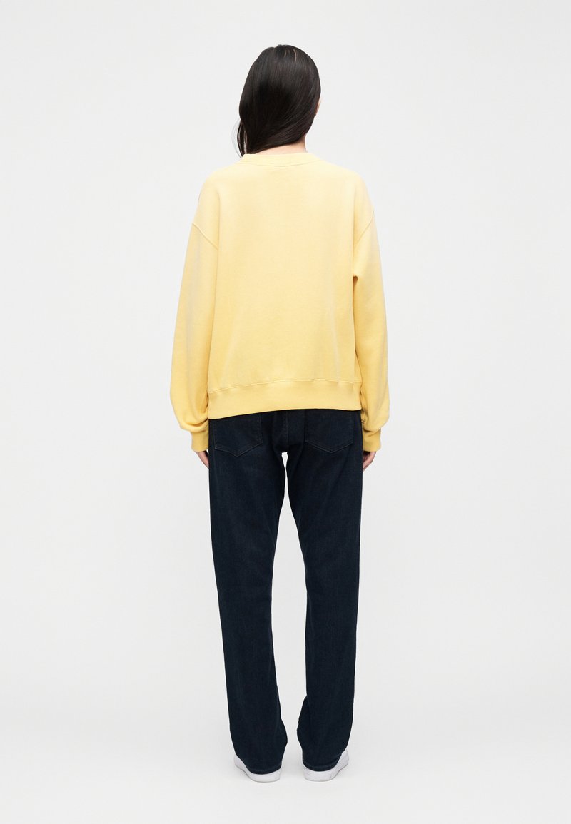Sweat-shirt en coton jaune avec épaules tombantes, poignets et ourlet côtelés, associé à un jean ample bleu foncé et des baskets blanches.
