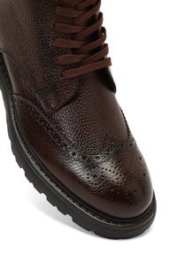 Ciemnobrązowy skórzany but z teksturowanym wykończeniem, z delikatnymi detalami w stylu brogue, płaskimi sznurowadłami i solidną czarną podeszwą.