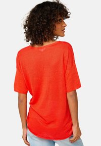 Red, loose-fitting top with short sleeves and a wide neckline. Made of lightweight fabric with subtle texture, featuring a rounded hem.