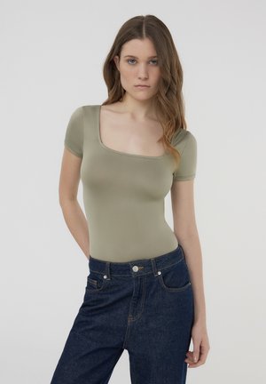 Body verde oliva con scollatura quadrata e maniche corte, abbinato a jeans denim blu scuro con tasche classiche e cuciture.