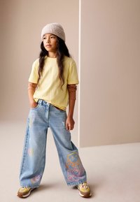 Ung person med langt hår iført en beige strikhue, lagdelte gule og brune overdele, vidde jeans med farverige smily-ansigter og gule sneakers.