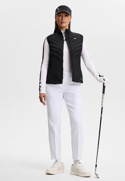 Kvinde iført sort vest, hvid skjorte med lange ærmer, hvide bukser og kasket, der holder en golfkølle i klar position mod en ensfarvet baggrund.