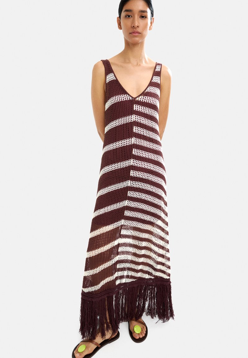 Szydełkowa maxi sukienka w burgundowe i białe paski, z dekoltem w kształcie litery V oraz frędzlami na dolnej krawędzi. Modelka nosi płaskie sandały z okrągłymi akcentami.