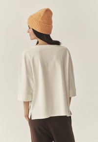 Biała oversize bluzka z rękawami o długości trzy czwarte i rozcięciami po bokach, zestawiona z pomarańczową, teksturowaną, dzierganą czapką. Prosty, minimalistyczny design.