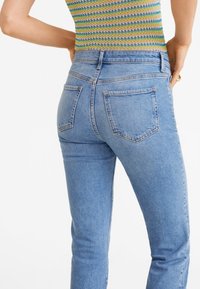 Persona vestida con jeans de tiro alto en azul y una camiseta sin mangas de rayas multicolores, con las manos en las caderas, mirando hacia el lado opuesto de la cámara.