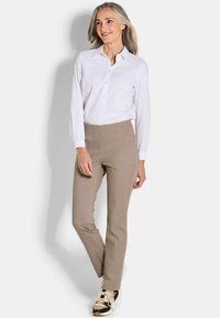 Vrouw met grijs haar, gekleed in een witte overhemd, beige broek en zwart-beige sneakers, staande tegen een witte achtergrond.