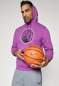 Sweatshirt roxo com capuz e um gráfico de basquetebol, usado com calções cinzentos; segurando uma bola de basquetebol laranja com um logotipo preto.