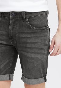 Nærbillede af en person iført rullede sorte denim shorts og en hvid skjorte, med højre hånd afslappet ved siden af.