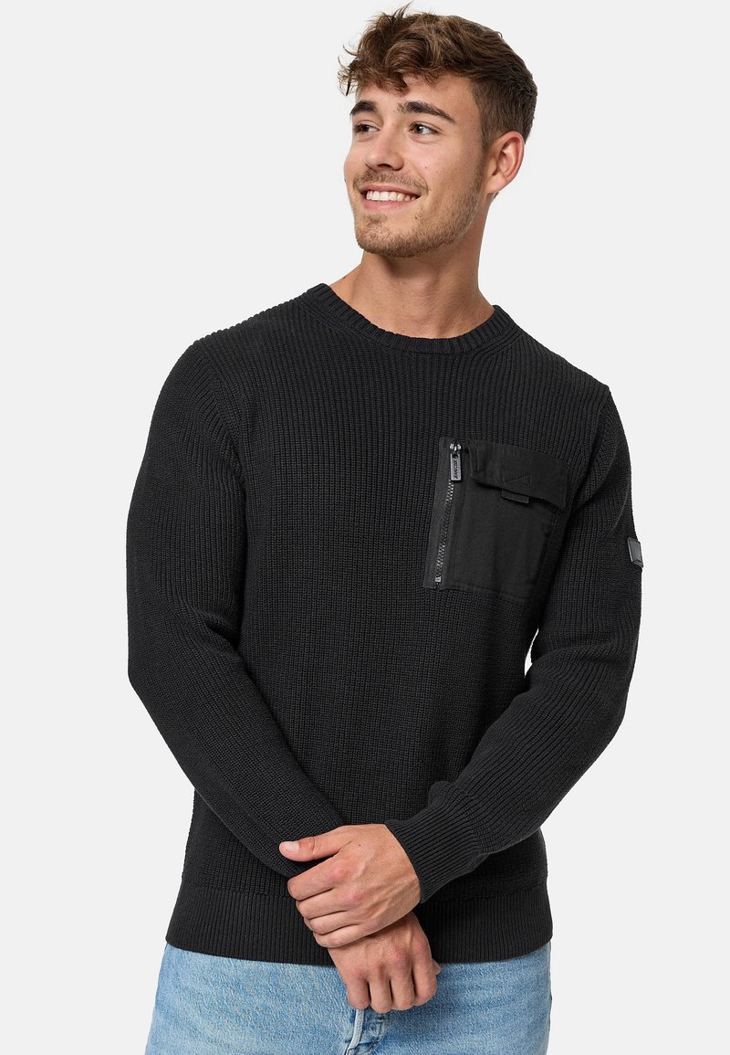 Pull en tricot noir à manches longues, avec un col rond et une poche zippée sur la poitrine. Il présente une texture côtelée et des poignets ajustés.