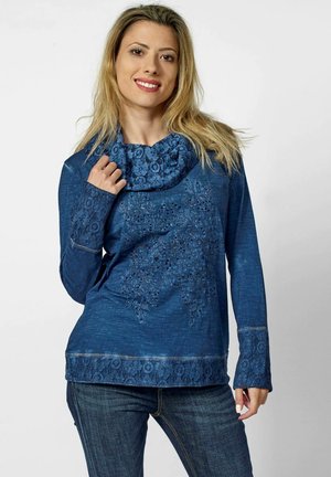 Haut à manches longues bleu présentant un col en cowl texturé et des accents en dentelle sur les manches et l'ourlet. Le matériau semble doux et tissé.
