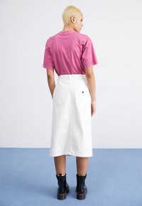 Biele denim midi sukňa s dvoma zadnými vreckami, kombinovaná s ružovým tričkom. Model nosí čierne členkové topánky so žltými akcentmi.