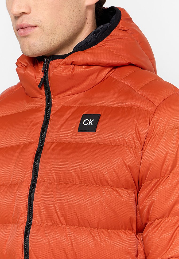 Chaqueta de plumas aislante de color naranja con capucha, que presenta un diseño acolchado, cierre frontal con cremallera y parche del logo negro de CK en el pecho.