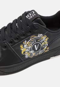 Svart sneaker laget av lærmateriale, med dekorativt hvitt og gull logo, rundt emblem, og teksturert gummisåle. Snøring.
