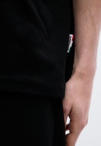 Μαύρο βαμβακερό μπλουζάκι με κοντά μανίκια, με εμφανές ετικέτα σε κόκκινο, λευκό και πράσινο στο τελείωμα, που κρατιέται κοντά σε χέρι.