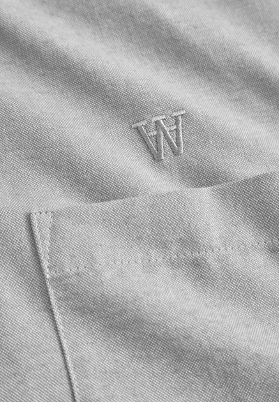 Un tissu gris clair avec une texture subtile présente un "W" brodé dans le coin supérieur gauche et une poche sur le côté droit.