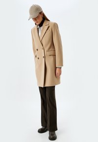 Abrigo beige de doble botonadura con solapas con muesca, dos bolsillos delanteros, llevado sobre un cuello de tortuga marrón, combinado con pantalones marrones y una gorra estampada.
