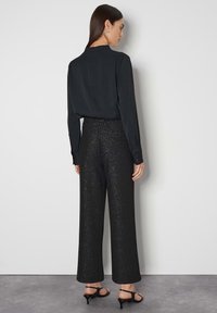 Zwarte blouse met lange mouwen, hoge hals en een ritssluiting op de rug. Hoge zwarte broek met een gestructureerde, glinsterende afwerking en wijde pijpen.