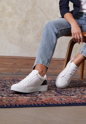 Persoon zittend op een houten stoel, gekleed in lichtblauwe spijkerbroek en witte sneakers met zwarte hieldetails, op een patroonrijke vloerkleed.