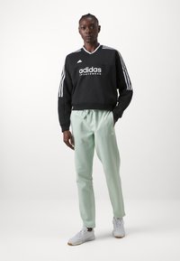 Sweatshirt preto com o logo "adidas" em branco e três riscas brancas nas mangas. Calças de fato de treino verde menta, combinadas com sapatos brancos e sola clara.
