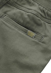 Tkanina w oliwkowozielonym kolorze o gładkiej fakturze, z tylną kieszenią i żółtą metką marki z napisem "KENT BRAND AUTHENTIC EST. 1982".