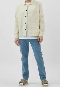 Veste beige clair avec des boutons noirs, deux poches avant ; assortie à un pantalon bleu clair et des sandales verdâtres sur des chaussettes blanches.
