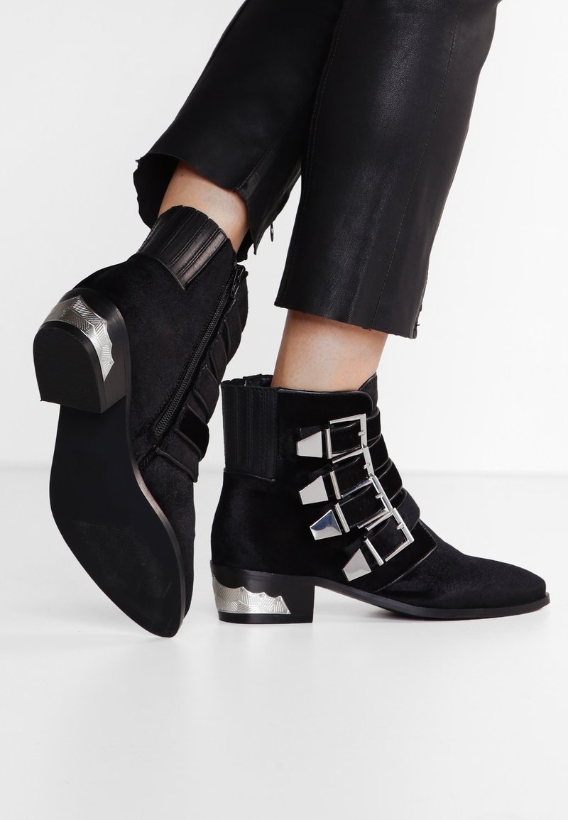 Stivaletti neri in suede con un tacco spesso decorato con accenti metallici argentati, dotati di tre cinturini con fibbie metalliche argentate e una chiusura con zip sul retro.