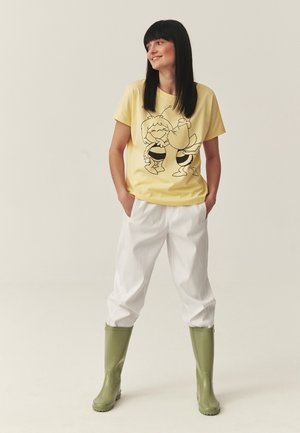 Kobieta o prostych czarnych włosach, ubrana w żółtą koszulkę z motywem kreskówkowej pszczoły, białe spodnie i zielone gumowe kalosze, stojąca z rękami w kieszeniach.
