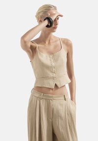Beige kort tanktopp med justerbara axelremmar, framsida med knappar och en texturerad tyg, kombinerad med lösa beige högmidjade byxor.
