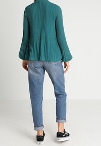 Blusa plissettata teal con maniche lunghe a campana, abbinata a jeans azzurri arrotolati e scarpe da ginnastica nere in tela. Sfondo bianco.