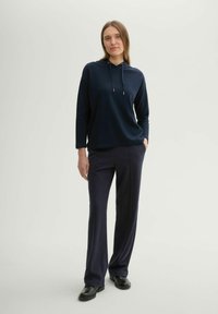 Felpa con cappuccio color blu navy in tessuto testurizzato, abbinata a pantaloni scuri su misura blu. Il modello sta in piedi con le mani nelle tasche, indossando scarpe nere.