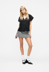 Ξανθιά γυναίκα με μαύρο κοντομάνικο τοπ, μίνι φούστα ασπρόμαυρος καρό, λευκές κάλτσες και μαύρα παπούτσια Mary Jane που στέκεται μπροστά σε λευκό φόντο.