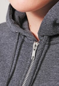 Felpa grigia con zip realizzata in un tessuto morbido, dotata di una cerniera argento e un cappuccio foderato con lacci. Vista ravvicinata del collo.