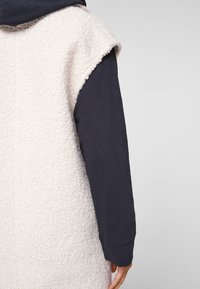 Gilet in pile color crema con superficie texturizzata, abbinato a una camicia a maniche lunghe blu navy sotto. Design semplice, senza chiusure visibili.