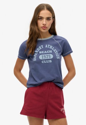T-shirt bleu à manches courtes avec du texte et des finitions bleu clair, présentant un col rond et une coupe décontractée. Associé à un short rouge à taille élastique.