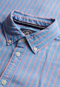 Chemise bleue à boutons avec rayures verticales roses et boutons de couleur crème, étalée à plat montrant le col et l'étiquette à l'intérieur.