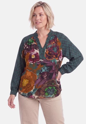 Vrouw draagt een blouse met bloemenpatroon met groene geometrische mouwen en een beige broek, staand met één hand op haar heup.